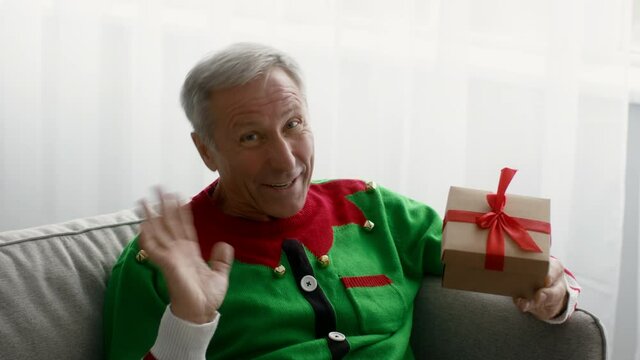 Funny senior man wearing christmas elf costume holding gift, waving at camera