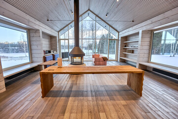 Country house living room interior with panoramic windows with table, sofa and stove, natural wood parquet and walls