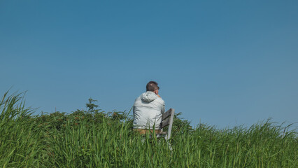 On a green meadow, among lush grass, there is a bench. A man in a windbreaker is sitting against a clear blue sky. View from the back. Kamchatka