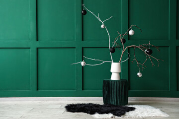 Tree branches with Christmas balls in vase on pouf near green wall