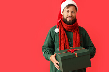 Handsome man in Santa hat with Christmas gift on red background