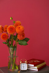Vase with beautiful dahlia flowers, alarm clock, books and eyeglasses on table against color background, closeup