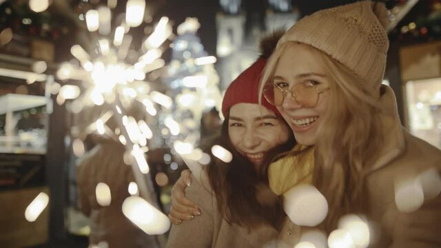 Adorable young girlfriends laughing and hugging at festive cozy fair in the central square of Prague. Gorgeous women burning blazing bengal lights and spending Christmas night together.