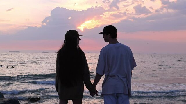 Young couple looking at each other and holding hands during sunset at the beach as waves roll in from the Pacific ocean.