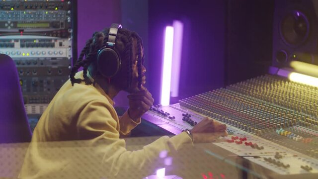 Young African American man in headphones using mixing console in music recording studio with neon light