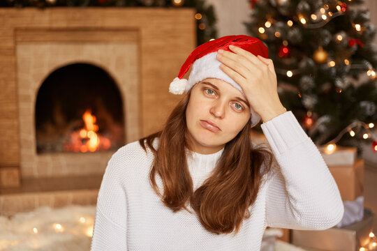 Unhealthy Caucasian woman wearing white sweater and santa claus hat, sitting near fireplace and xmas tree and keeping hand on her forehead, suffering headache, flu symptoms.