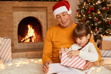 Fototapeta na wymiar Kind senior man wearing orange sweater and santa claus hat posing with little girl opening his Christmas present with surprised facial expression. Merry xmas and happy new year.