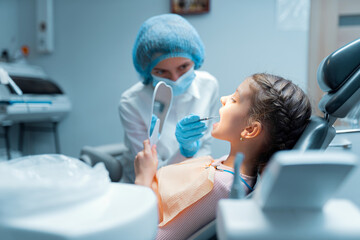 Little girl patient and female dentist are checking teeth at mirror after treatment in dental office