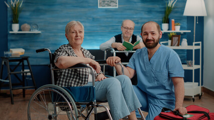 Portrait of old disabled woman and man nurse looking at camera in nursing home. Retired patient with chronic issues sitting in wheelchair while medical assistant preparing for examination