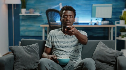 POV of adult switching channels with TV remote control. Person watching television and changing channel while having popcorn snack for free time entertainment. Man looking at camera