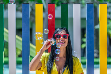 Latina woman smiling and blowing bubbles soap in a yellow dress against a background of a pool and...