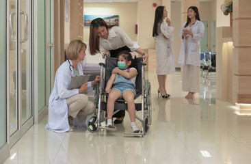 Female doctor kneeling and talking with little girl in wheelchair with her mother in hospital hall.