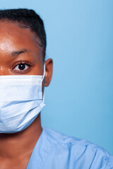 Closeup of african american specialist nurse with protective medical face mask to prevent infection with covid19 working at illness expertise in studio. Portrait of therapist woman in blue backgroud