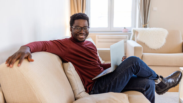 Portrait of smiling African American man in glasses sit at sofa in office working on laptop, happy biracial male worker look at camera posing, busy using modern computer gadget at workplace - Powered by Adobe