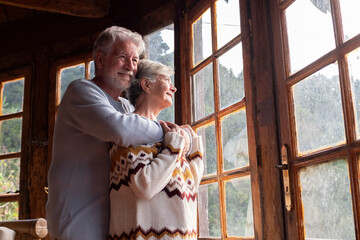 Elderly age relationship and love concept with couple of mature man and woman hugging and looking outside the windows at home with happiness and joy together