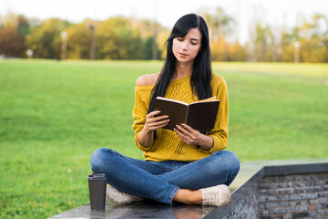 A beautiful and attractive Caucasian brunette girl in a yellow sweater is reading a book while sitting in the park.