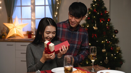 hide and seek time for presents. Loving asian husband closing wife eyes with hand and prepared Christmas surprise. cheerful girl laughing getting gift during xmas dinner time
