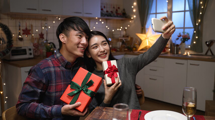 Excited young couple sitting at dinner table in kitchen exchanging with gift boxes during date night on valentines day. two lovers holding present and cellphone making selfie together