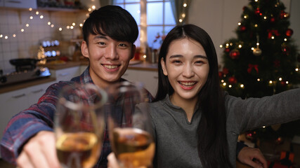 Two young woman and man looking at camera holding white wine glasses and toast. sweet couple smiling clinking champagne in video call for Christmas and New Year at home kitchen by xmas tree