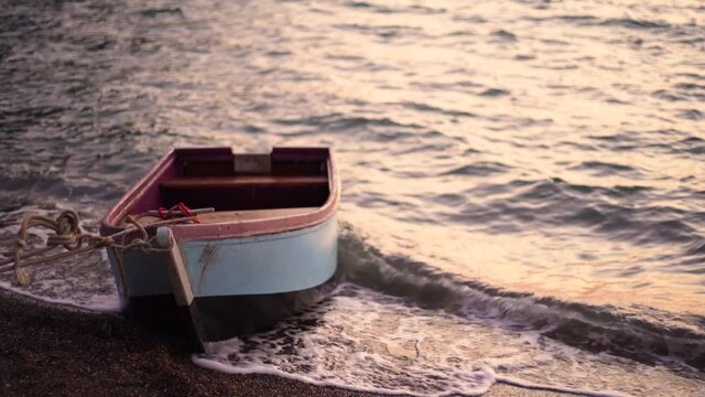 Close-up view of small wooden boat floating on waves of golden sunset sea water on empty sunset evening beach