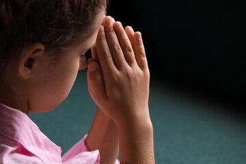 Cute little girl with hands clasped together praying on blurred background, closeup