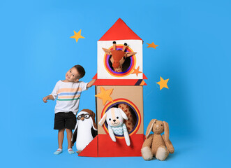 Cute little boy playing with cardboard rocket and soft toys on light blue background