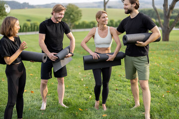 Group of pleased european people folding fitness mats after training on green meadow. Concept of healthy lifestyle. Idea of relaxation. Athletic men and women barefoot and wearing sportswear