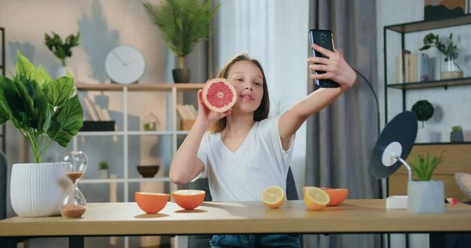 Beautiful happy carefree smiling teen girl sitting at the table in cozy sitting room and making selfie with grapefruit on her smartphone