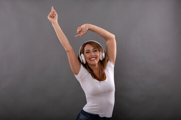 Cheerful and happy attractive brunette student girl with delighted smile dressed in white shirt enjoys in music on big white headphones isolated on gray background