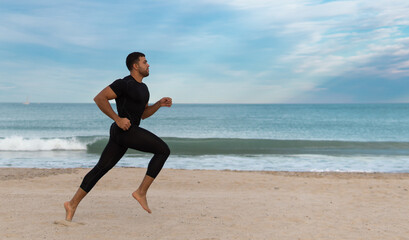 hombre moreno y fuerte de cuerpo atlético indio o pakistaní practica deporte y corre con ropa ajustada negra en un playa de arena con un fondo azul 