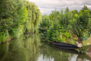 One of the navigable canals of the picturesque marshlands of the Regional Natural Park of the Marais Poitevin, with rowboats and weeping willows on the water - obrazy, fototapety, plakaty