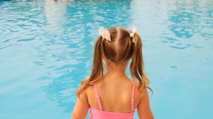 joyful little girl 4-5 years old, with ponytails, dressed in a pink swimsuit, looks at the blue water in the pool or in the sea on summer vacation, back view