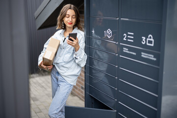 Young caucasian woman with parcel near automatic post terminal. Smiling girl holding smartphone and looking away on city street. Concept of smart delivery. Idea of modern shipping and logistics