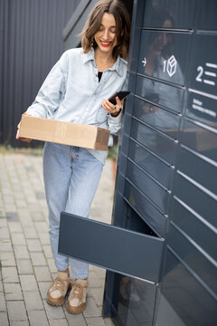 Young caucasian woman with parcel near automatic post terminal. Smiling girl holding smartphone and looking away on city street. Concept of smart delivery. Idea of modern shipping and logistics