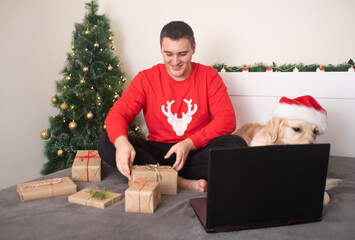 A man with a dog wearing a santa claus hat looks at a laptop screen. Christmas video greetings on social networks. New Year's home holiday. Happy New Year and Merry Christmas © deine_liebe