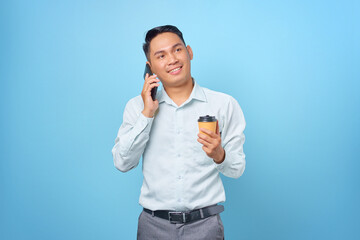 Smiling young handsome businessman talking on smartphone and hold a cup of coffee on blue background