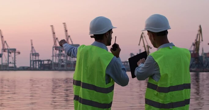 Men in helmets and reflective vests stand by seaside and regulate work of portal cranes via tablet and walkie-talkie during sunset