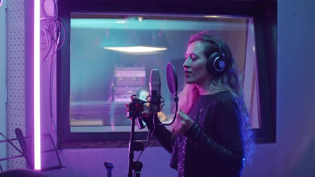 Beautiful female singer in headphones and shiny dress singing in microphone while recording new song in audio production studio