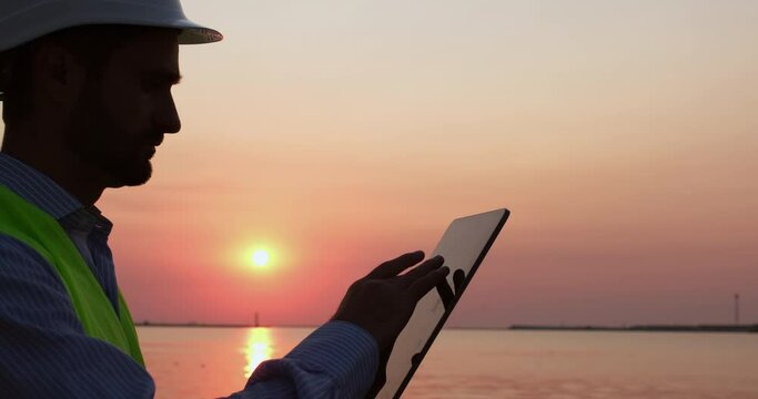 Port worker silhouette in helmet and vest controls movement of ships and cargo cranes in port using tablet PC at sunset closeup