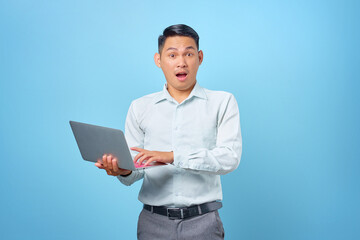 Naklejka na ściany i meble Shocked young handsome businessman holding a laptop and looking at camera on blue background