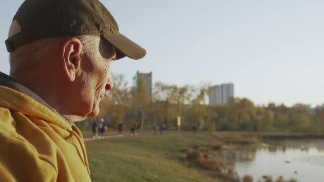 Lonely elderly grandfather in cap stands at autumn city park and looks at walking people near small lake or pond. Oldness and life style concept.