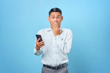 Fototapeta premium Shocked young handsome businessman holding smartphone and covering mouth on blue background