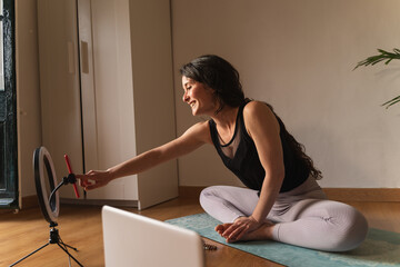 Happy young woman wearing sports clothes sitting at home and recording her yoga workout.