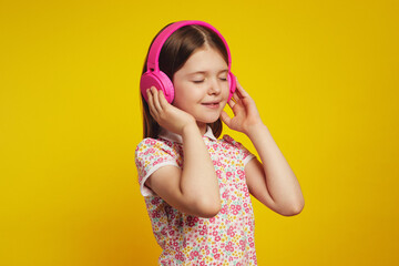 Obraz na płótnie Canvas Attractive kid girl in stylish summer clothes, listening to music in pink headphones with closed eyes, isolated over yellow studio background