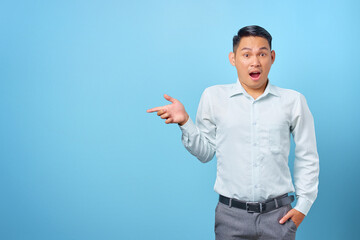 Fototapeta premium Shocked young handsome businessman pointing finger away at copy space on blue background