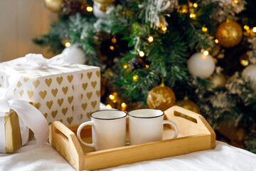 Gift box, wooden tray and two white mugs on the bed on a white sheet, near a decorated Christmas tree. Cozy morning. Coffee in bed