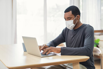Black businessman in wears medical protective mask sitting at a table at home on laptop computer. Self-isolation quarantine, new normal. Social Distancing At Work During Coronavirus Covid-19 Pandemic