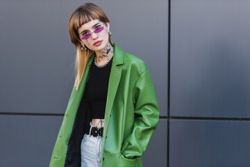 tattooed woman with piercing looking at camera while posing with hand in pocket of green jacket