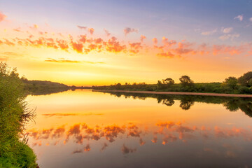 Fototapeta na wymiar Scenic view at beautiful summer river sunset with reflection on water with green bushes, grass, calm water ,deep cloudy sky and glow on a background, spring , evening landscape
