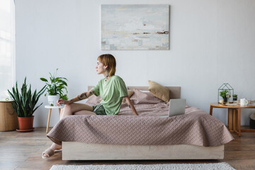 young tattooed woman sitting on bed near laptop in modern bedroom